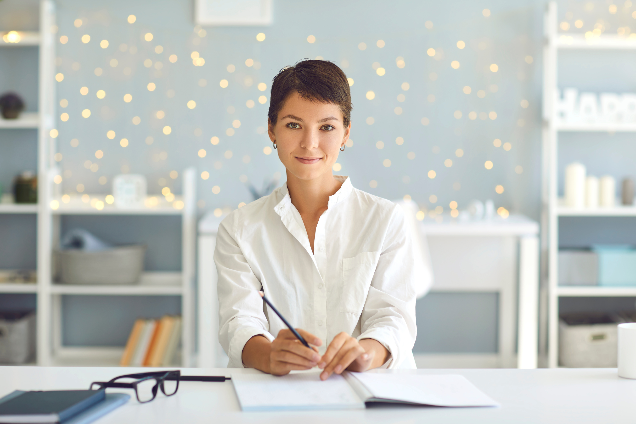 Confident Woman Sitting at Desk with Pen and Open Notebook, Looking at Camera as If Recording Video